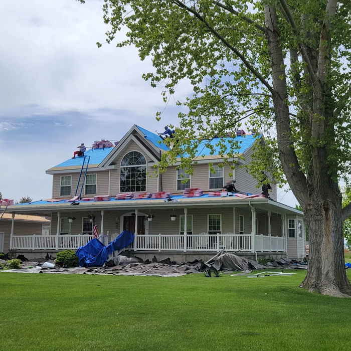 Storm-damaged two-story house with blue tarp on roof, broken front porch, and debris in the yard of idaho falls home