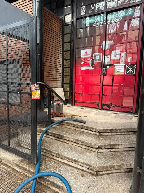 Fachada roja con carteles, escalones de piedra y una manguera azul junto a una verja metálica.