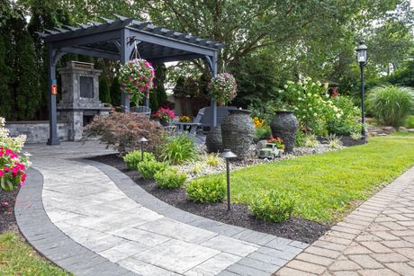 Landscaped garden with stone patio, curved walkway, and lush flower beds beside a pergola. Landscaped garden with stone patio, curved walkway, and lush flower beds beside a pergola.