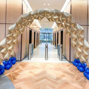 Gold balloon arch framing an office hallway, with blue balloons along the floor and wood herringbone flooring.