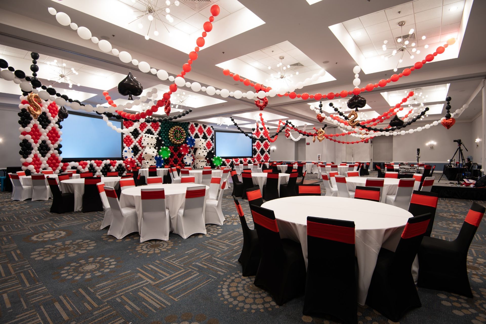 Red, black and white linking balloon strands with suspended casino accents across the event room ceiling.