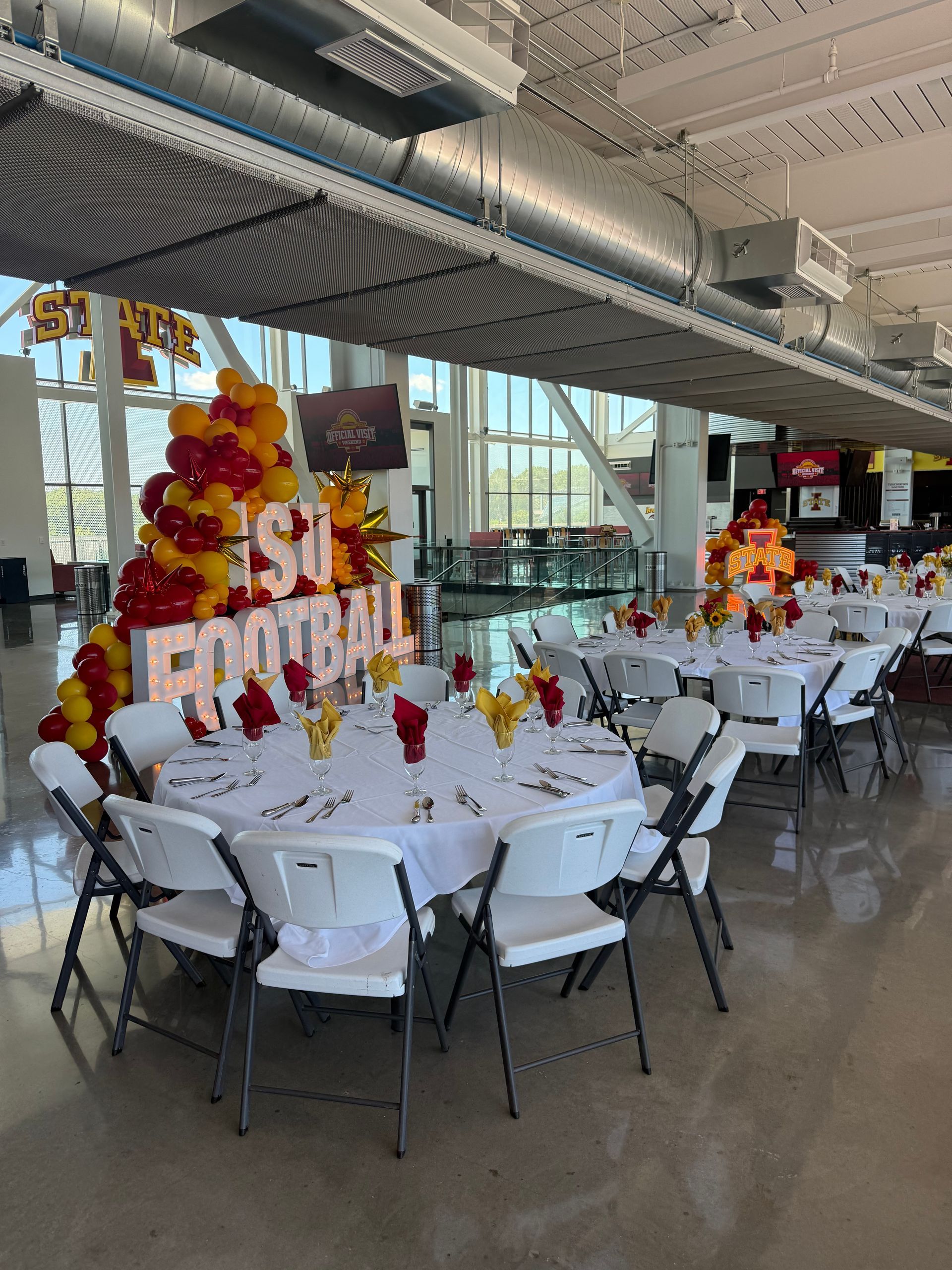 Decorated banquet tables in an industrial hall with red, gold, and white balloons and a “She’s a bomb” sign