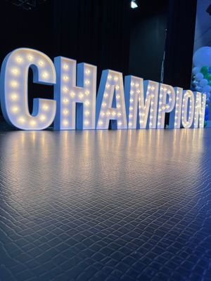 Large illuminated “CHAMPION” letters on a stage with a reflective floor and dark blue lighting.