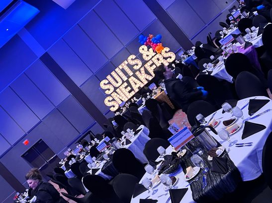 Banquet hall with round tables under blue lighting, featuring a lit “Suits & Sneakers” sign on stage