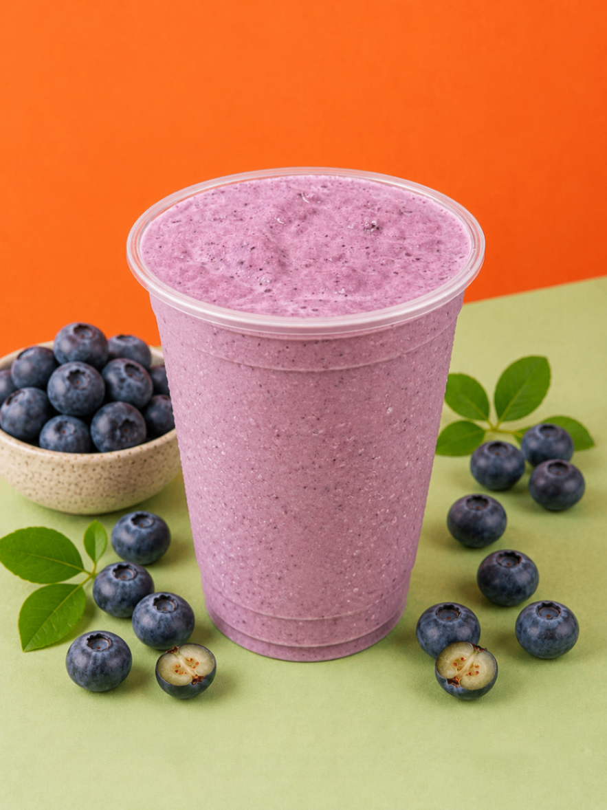 Purple blueberry smoothie in a clear cup, surrounded by fresh blueberries on a green and orange background