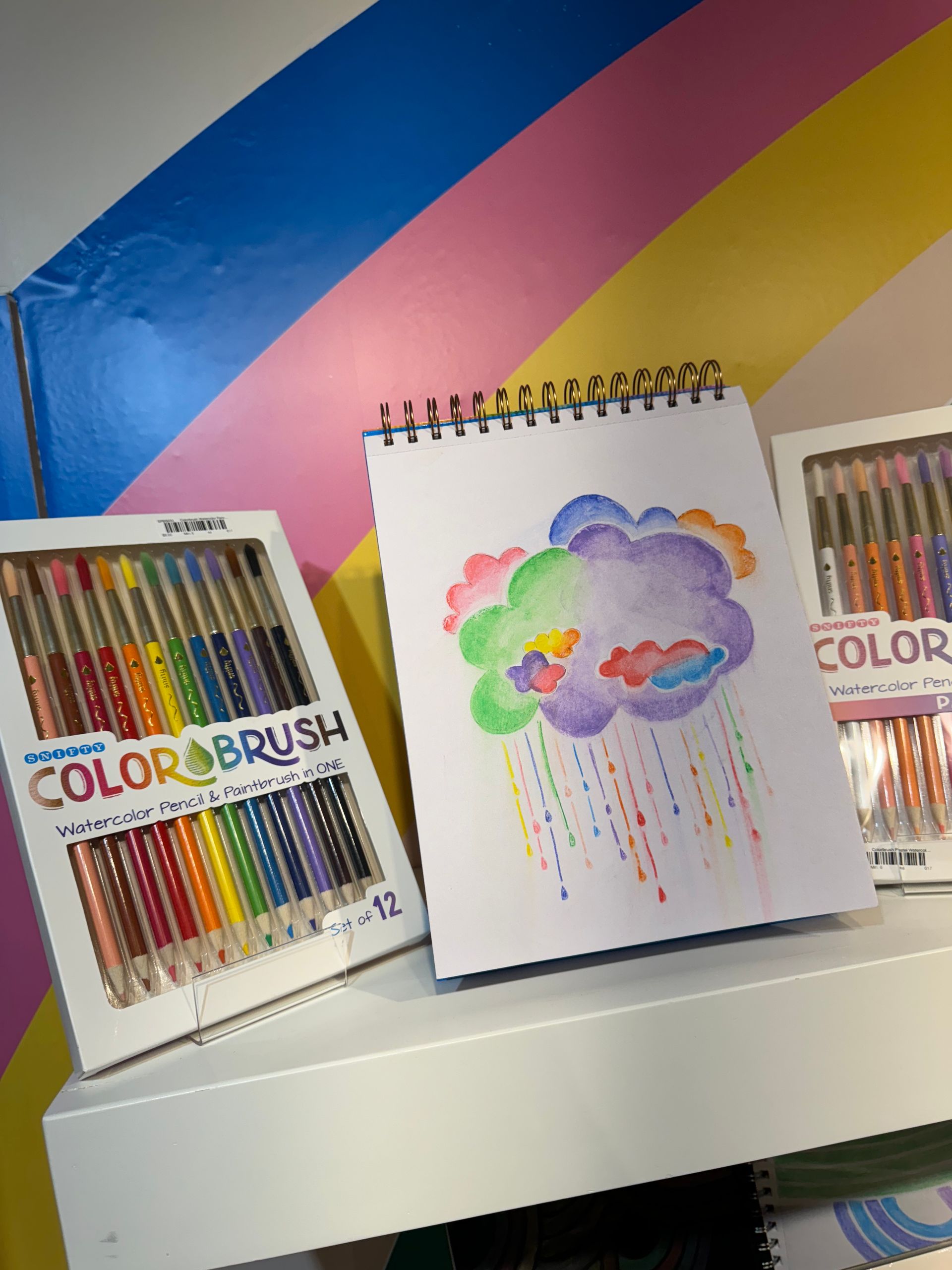 A box of color brush colored pencils sits next to a drawing of a cloud