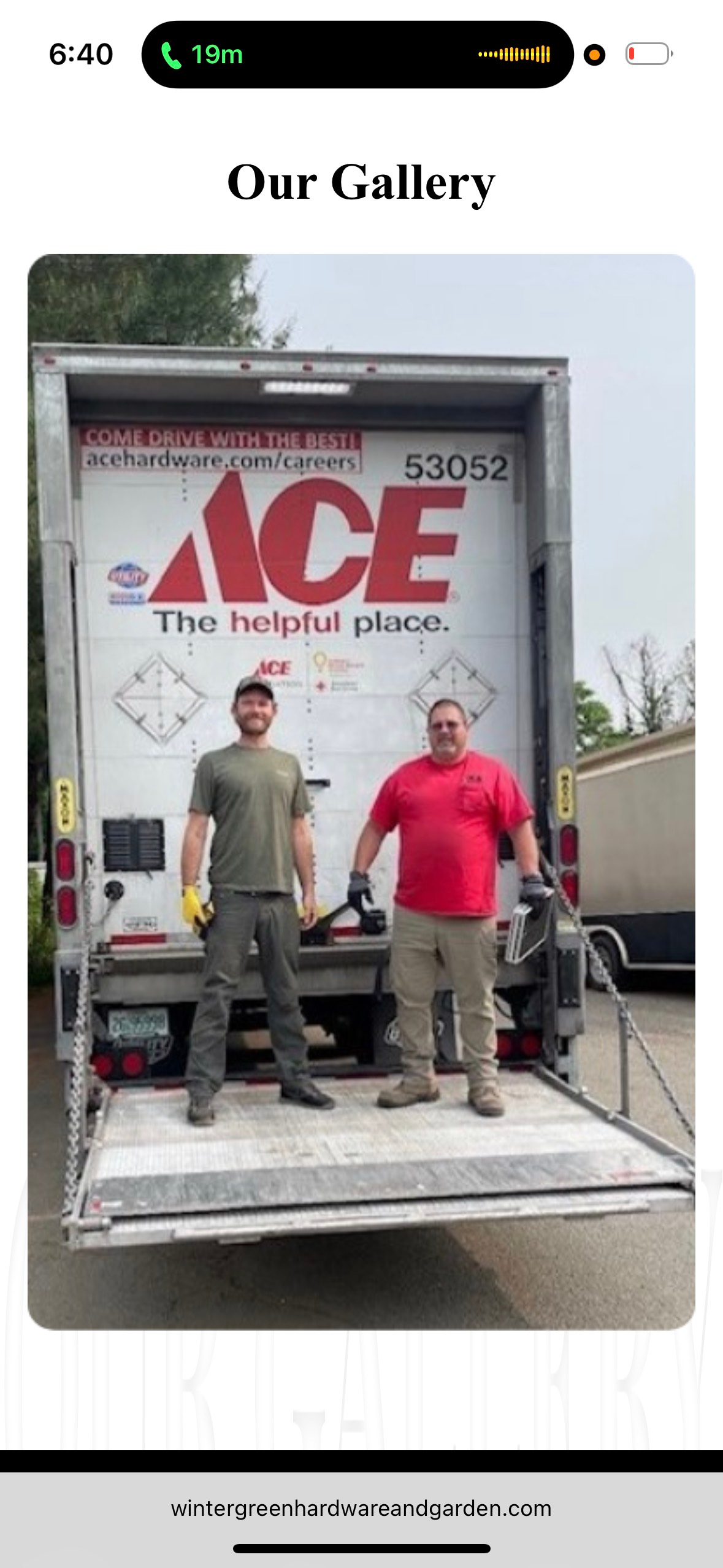 Two men stand on the back of an Ace Hardware truck. The men are holding tools and are wearing work clothes.