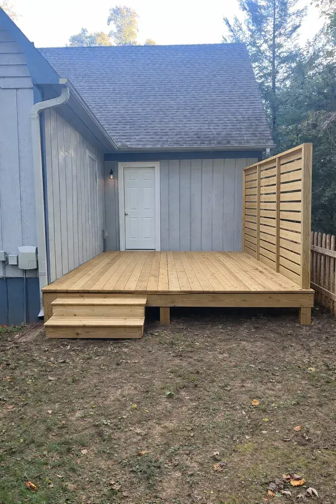 Wooden backyard deck with steps and privacy screen beside a gray house, surrounded by trees.