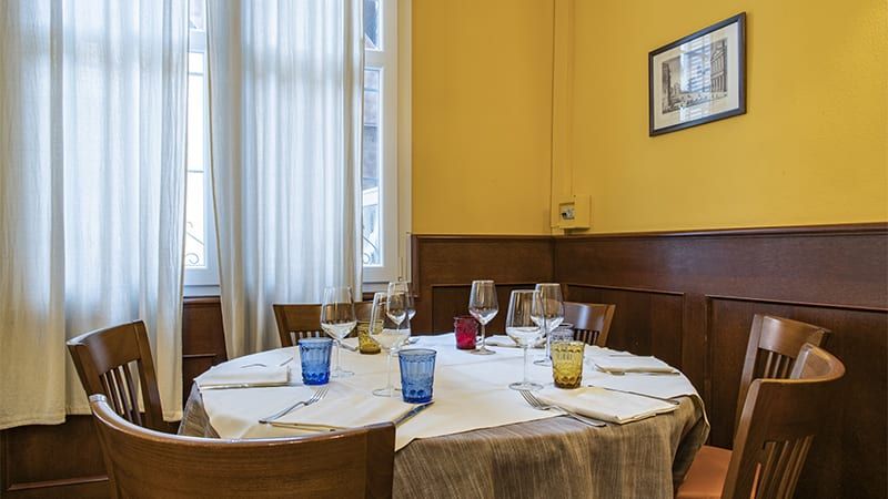 Tavolo da pranzo rotondo apparecchiato con bicchieri e sedie, vicino a una finestra con tende in un ristorante.