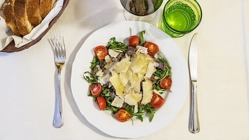 Insalata con pomodori e formaggio su un piatto bianco, servito con posate, pane e bicchiere d'acqua.
