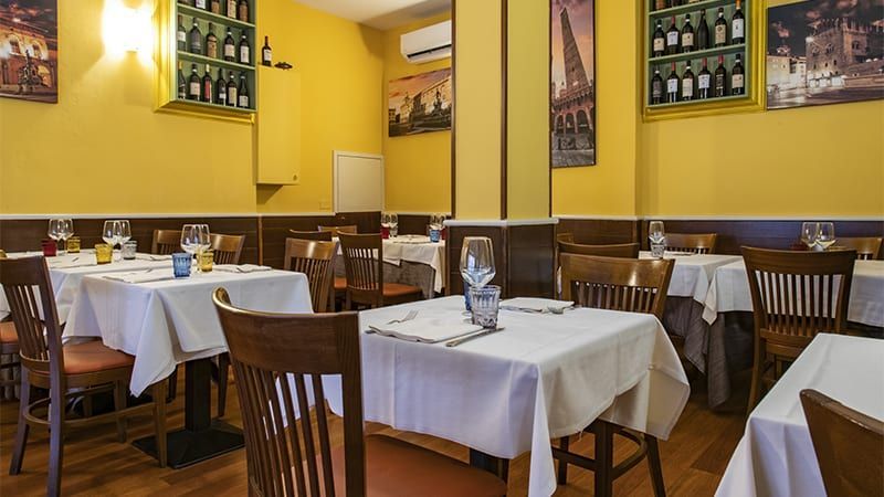 Interno del ristorante con tavoli apparecchiati per cenare; pareti gialle, sedie in legno e bottiglie di vino esposte.