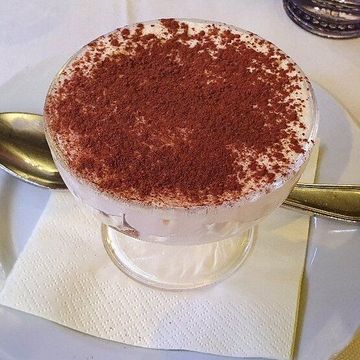 Dessert tiramisù servito in un bicchiere trasparente, spolverato con cacao in polvere, su un piatto bianco con tovagliolo e cucchiaio.