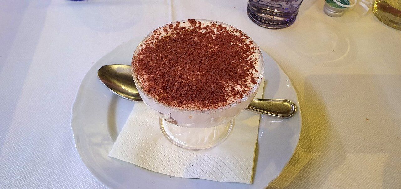 Dessert tiramisù in una ciotola di vetro su un piatto bianco, servito con un cucchiaio e un tovagliolo.