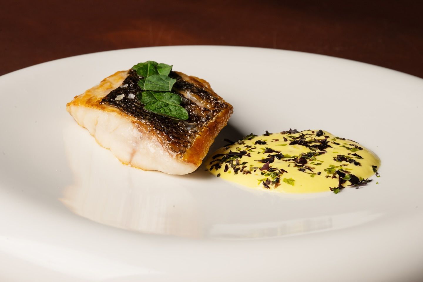 Pesce scottato con pelle dorata, erbe fresche e salsa gialla su un piatto bianco.