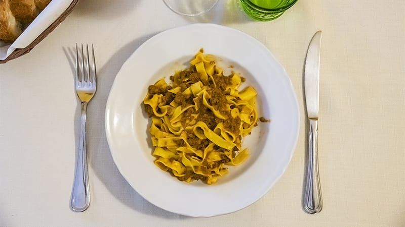 Piatto di pasta al ragù, servito con forchetta e coltello su una tovaglia bianca.