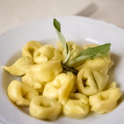 Piatto di tortellini, guarnito con salvia, in una ciotola bianca, su uno sfondo sfocato.