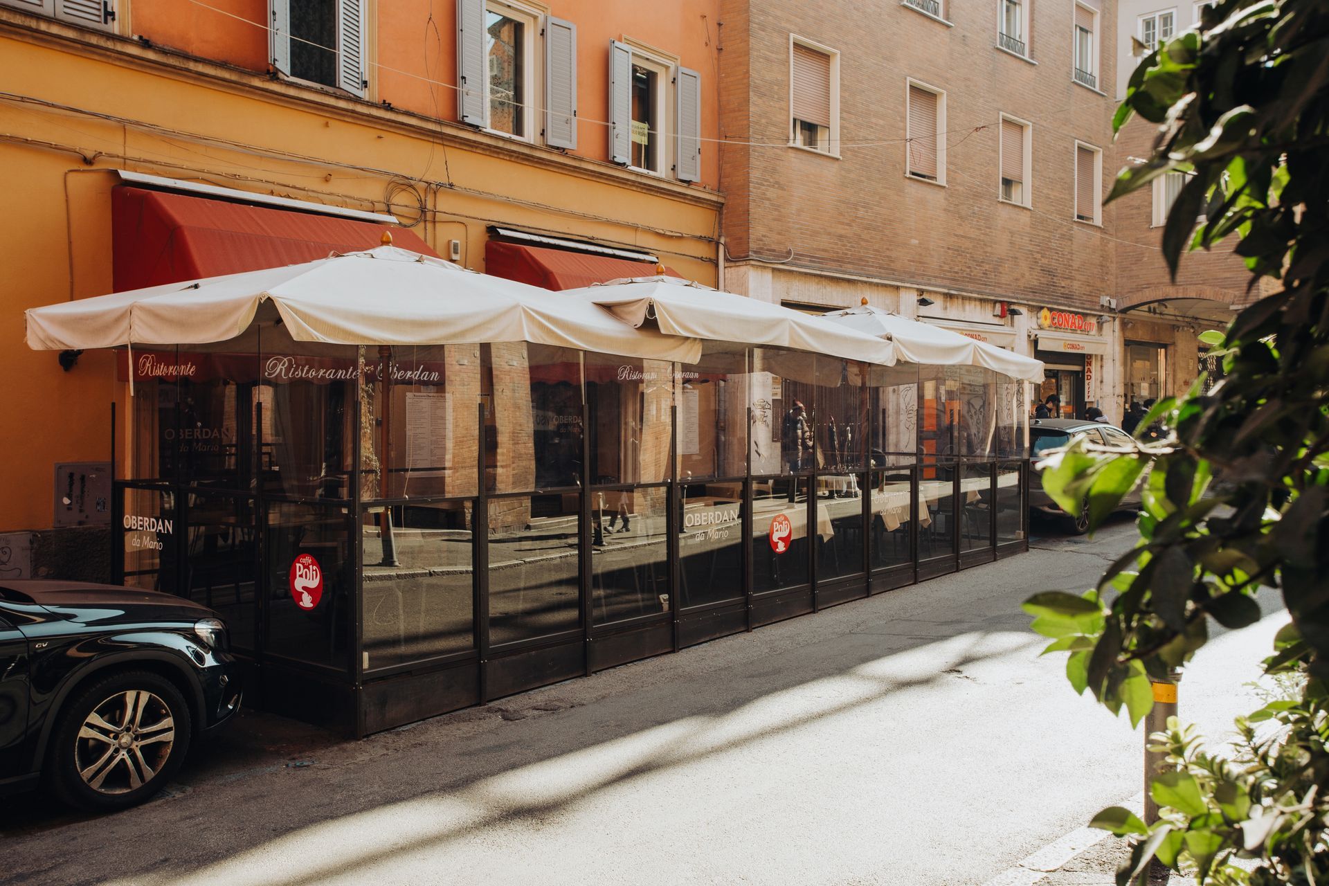 Area pranzo all'aperto con transenne trasparenti e ombrelloni bianchi lungo la strada.