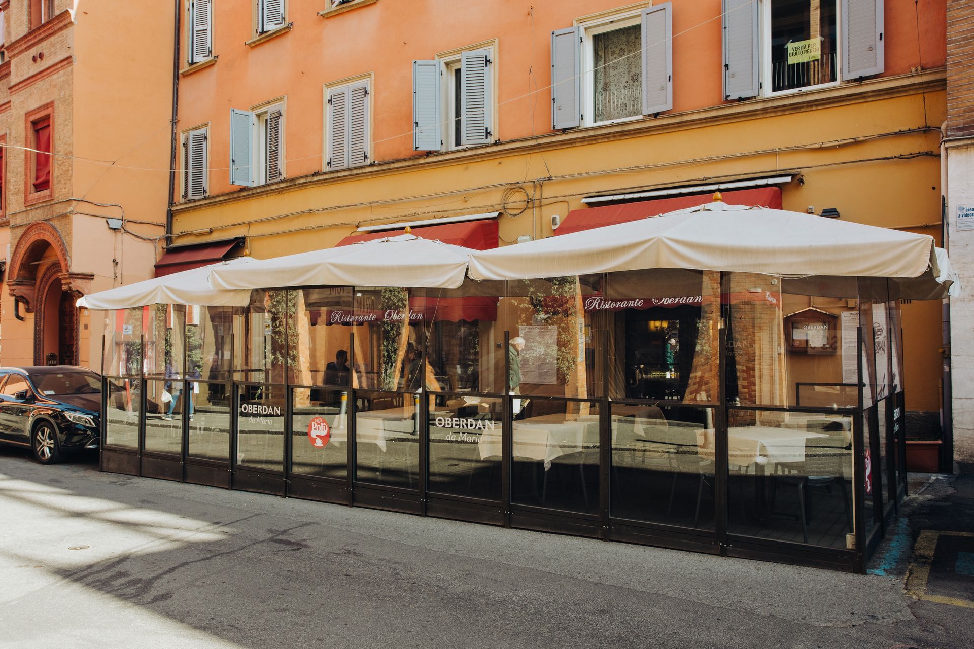 Esterno del ristorante con posti a sedere all'aperto su una strada cittadina; pareti di plastica trasparente, ombrelloni, tende da sole color marrone chiaro e rosso.