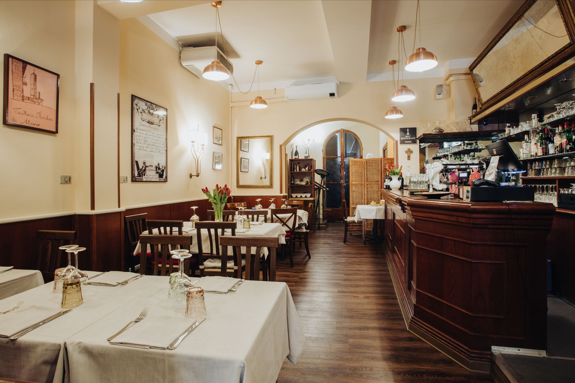 Interno di un ristorante con tavoli, bar e opere d'arte alle pareti. Luci calde, pavimenti e tavoli in legno.