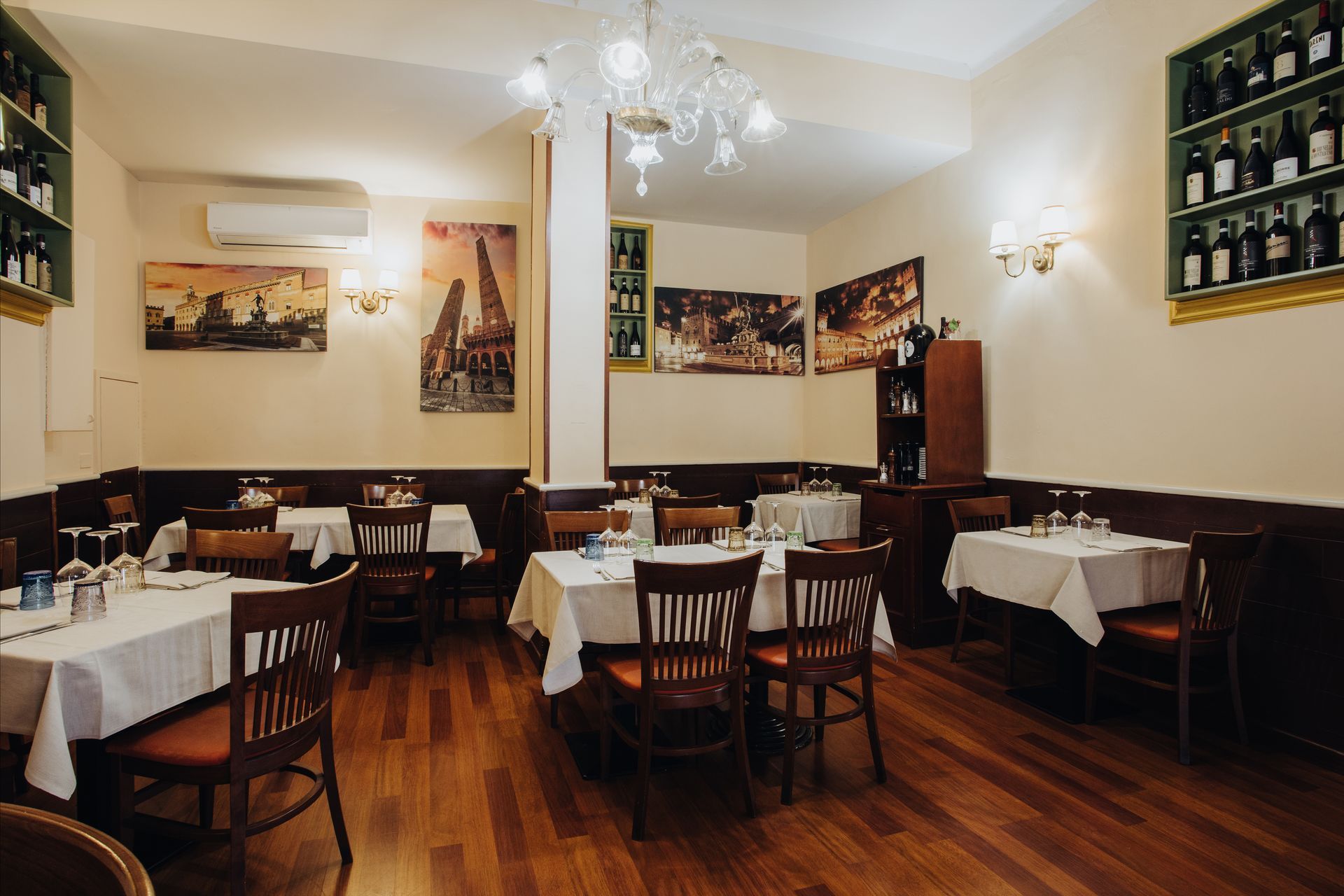 Interno di un ristorante italiano con pavimenti in legno, tovaglie bianche ed espositori per bottiglie di vino.