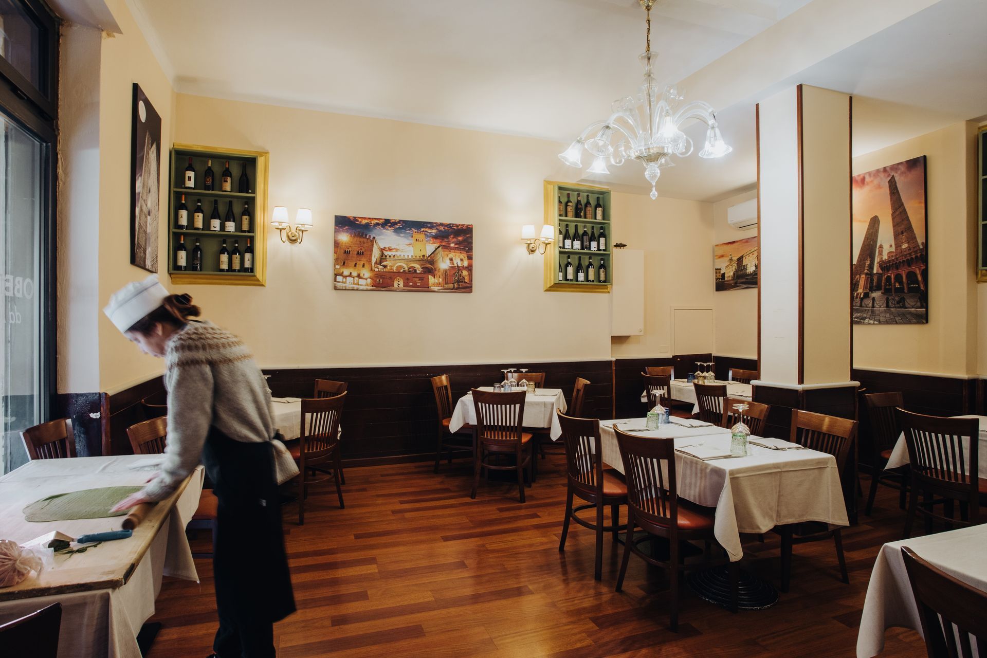 Interno di un ristorante con una persona che apparecchia un tavolo. Pavimenti in legno, tavoli con tovaglie bianche, opere d'arte alle pareti.