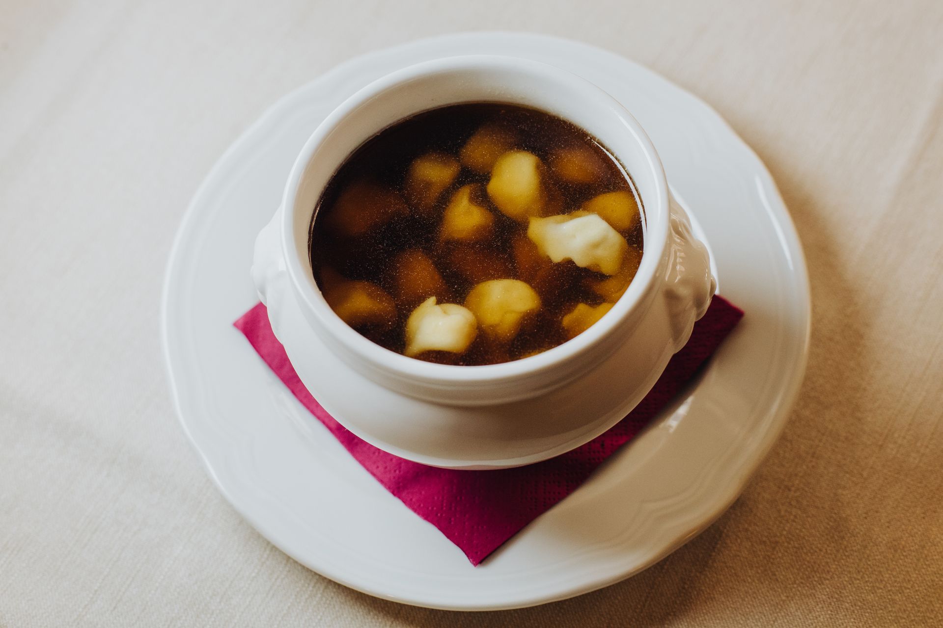 Zuppa in una ciotola bianca, appoggiata su un piatto bianco con un tovagliolo rosa sotto. La zuppa ha dei ravioli galleggianti.