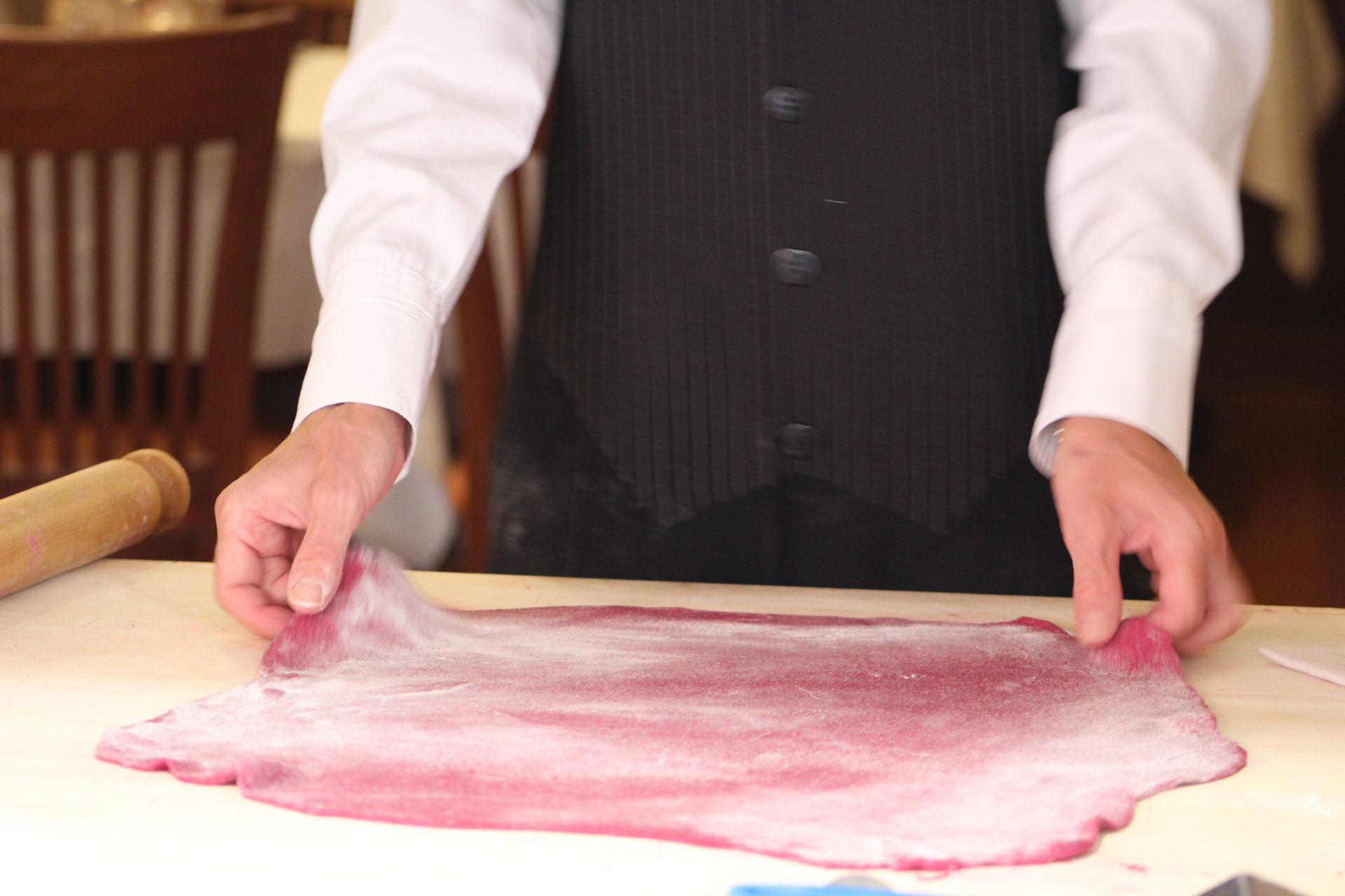 Persona in canottiera che stende un foglio di pasta rosa su una superficie bianca, con il mattarello nelle vicinanze.