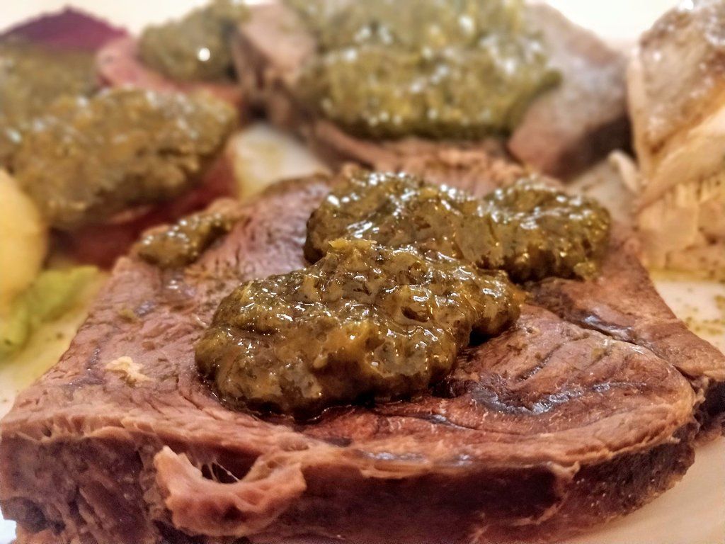 Carne cotta a fette condita con salsa verde. Primo piano con altri alimenti sullo sfondo.