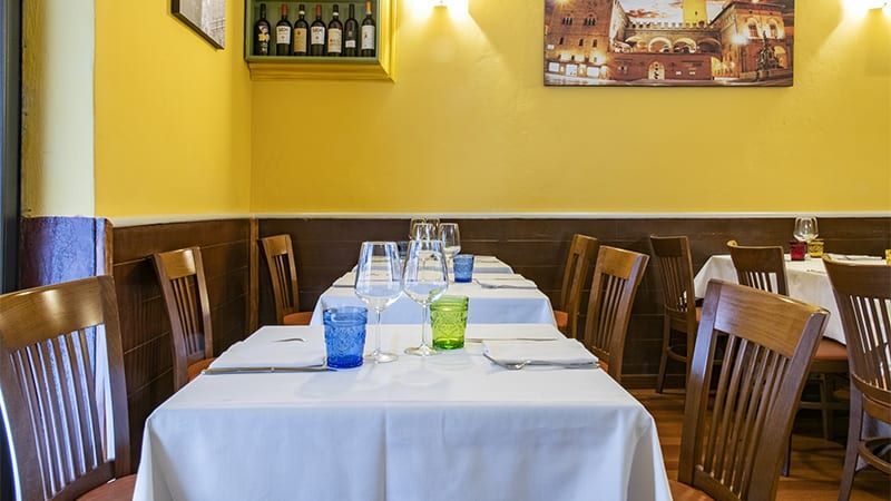 Interno del ristorante: tavoli apparecchiati con bicchieri e tovaglie, sedie in legno, pareti gialle, opere d'arte incorniciate.