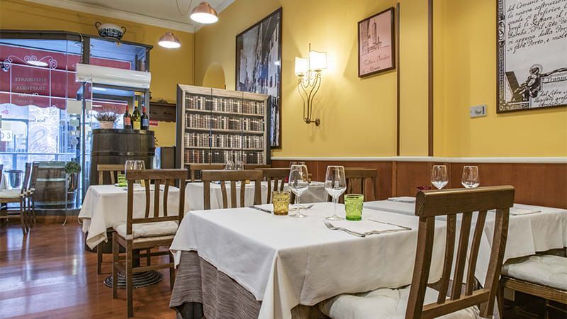 Interno di un ristorante con tavoli apparecchiati per cenare; pareti gialle, sedie in legno e uno scaffale da biblioteca.