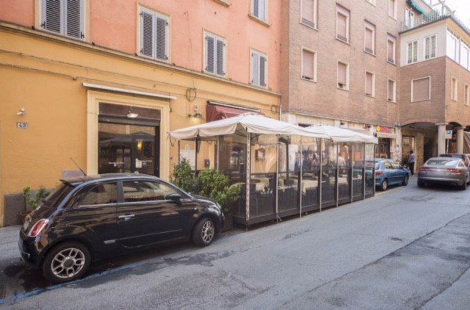 Auto nera parcheggiata in strada accanto ai tavoli all'aperto di un ristorante con recinzioni trasparenti. Edifici sullo sfondo.
