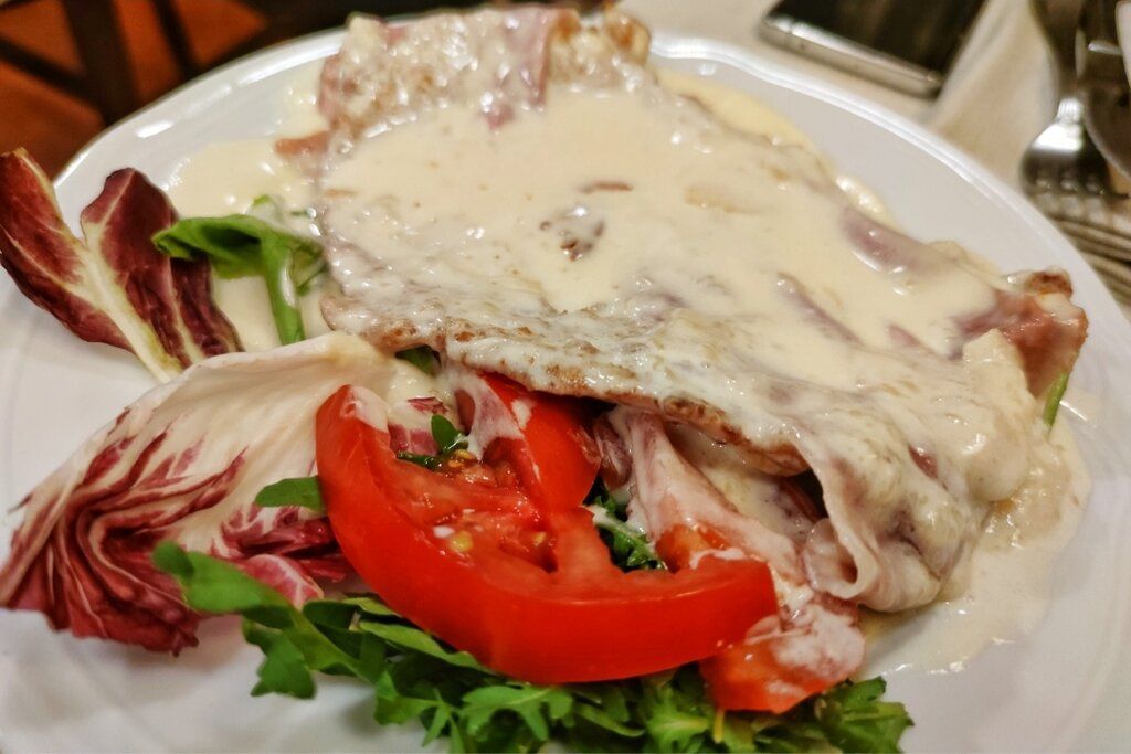 Piatto di pollo con salsa cremosa, prosciutto, fette di pomodoro e verdure su un piatto bianco.