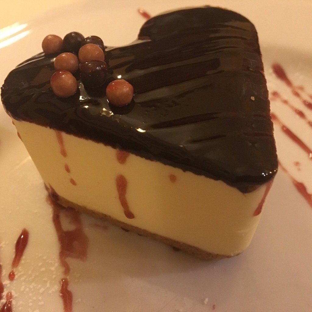 Cheesecake a forma di cuore con glassa al cioccolato e frutti di bosco su un piatto bianco.