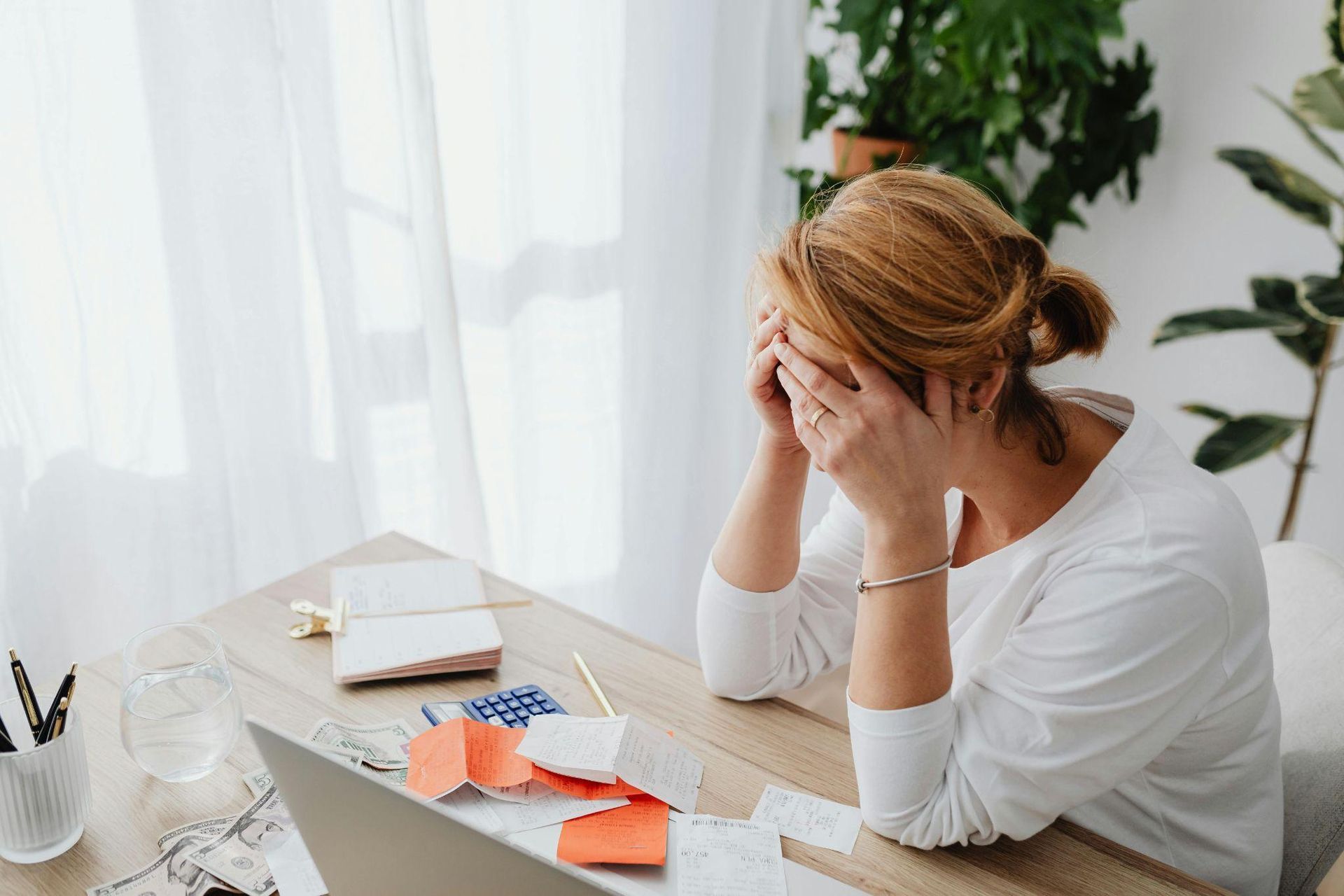 Woman stressed at a desk, hands on face; bills, calculator, laptop, and plants visible.