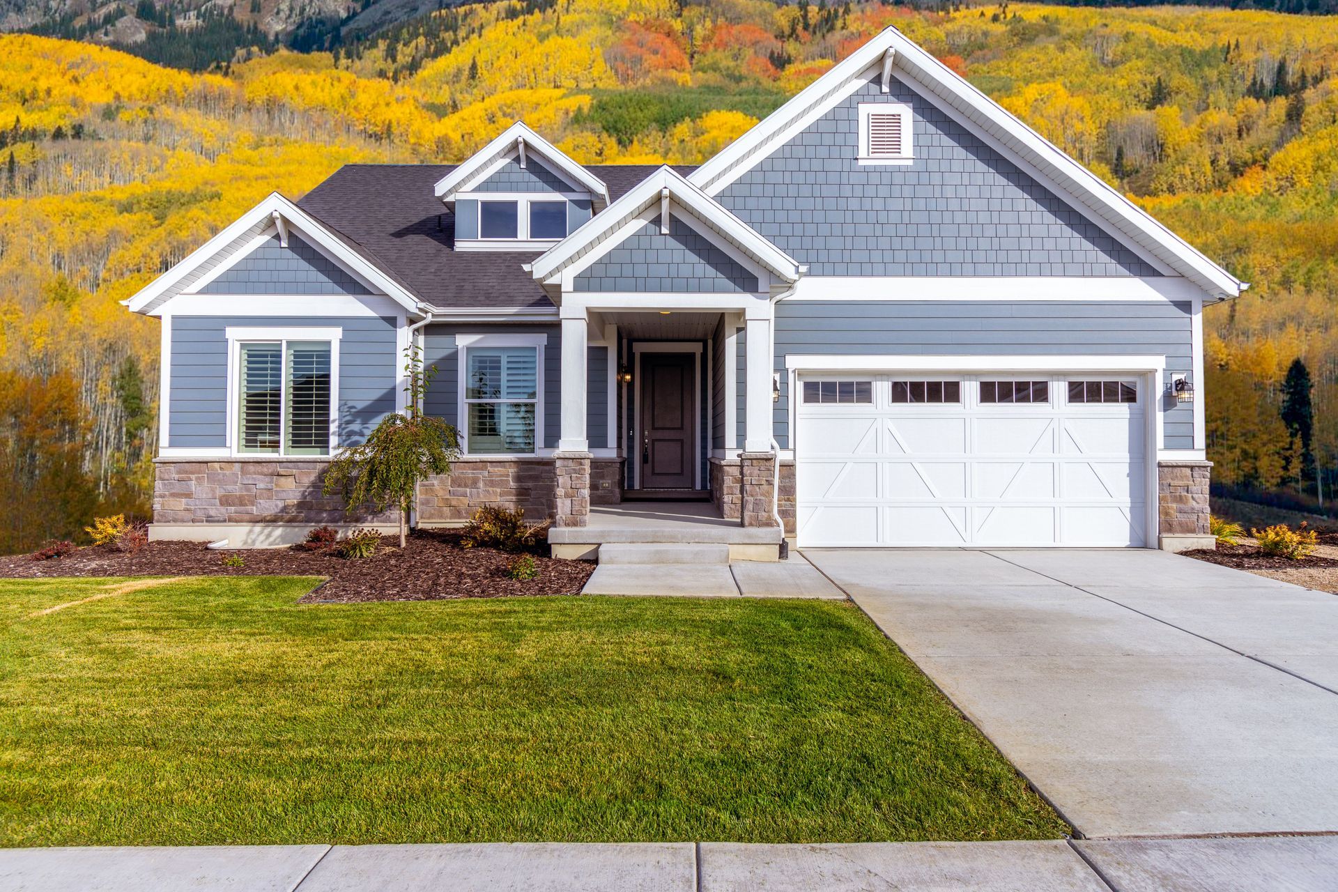Gray suburban house with a white garage, front lawn, and autumn trees in the background