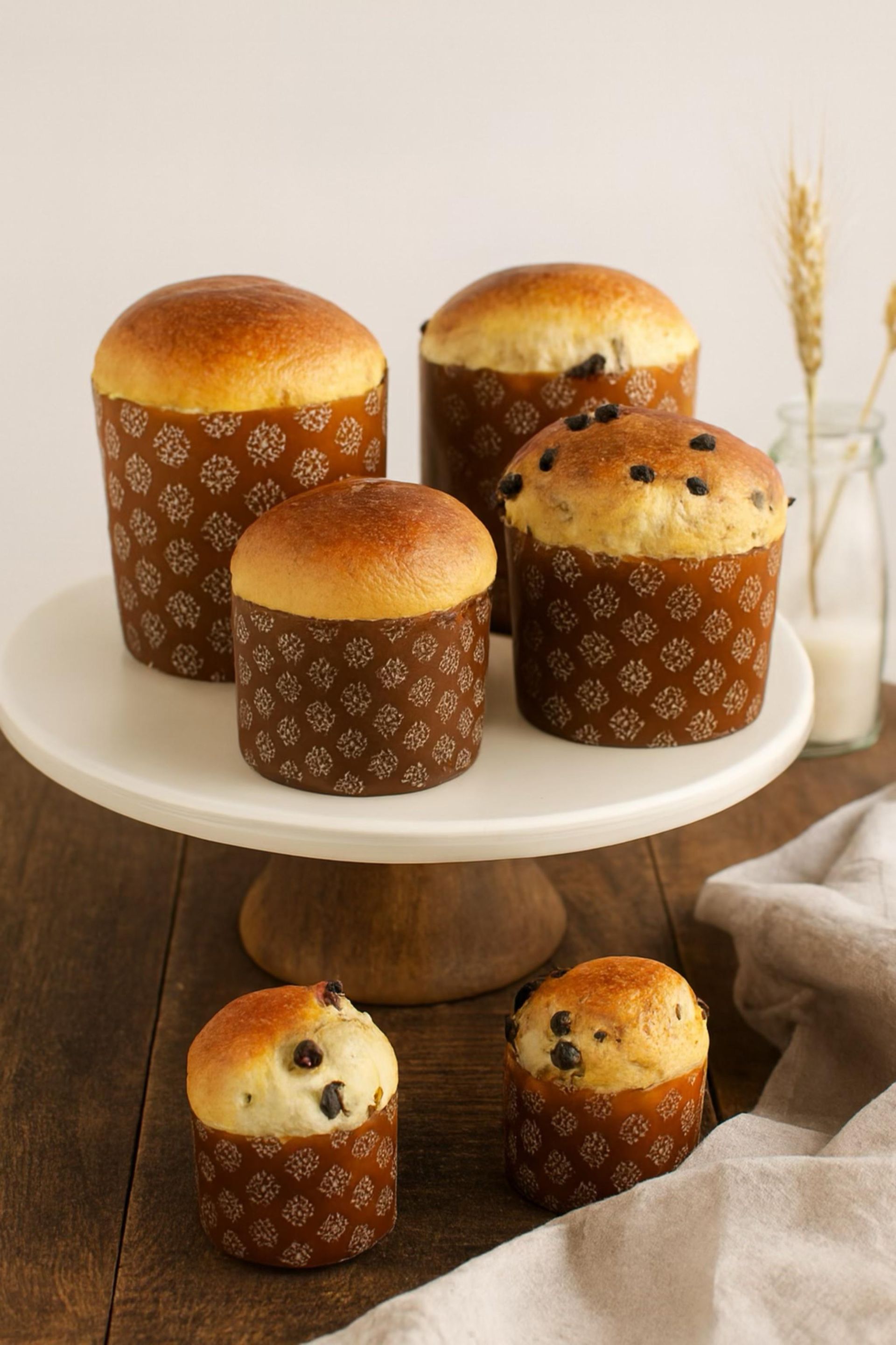 Surtido de panes dulces con corteza dorada, expuestos en un soporte para pasteles blanco y una mesa de madera.