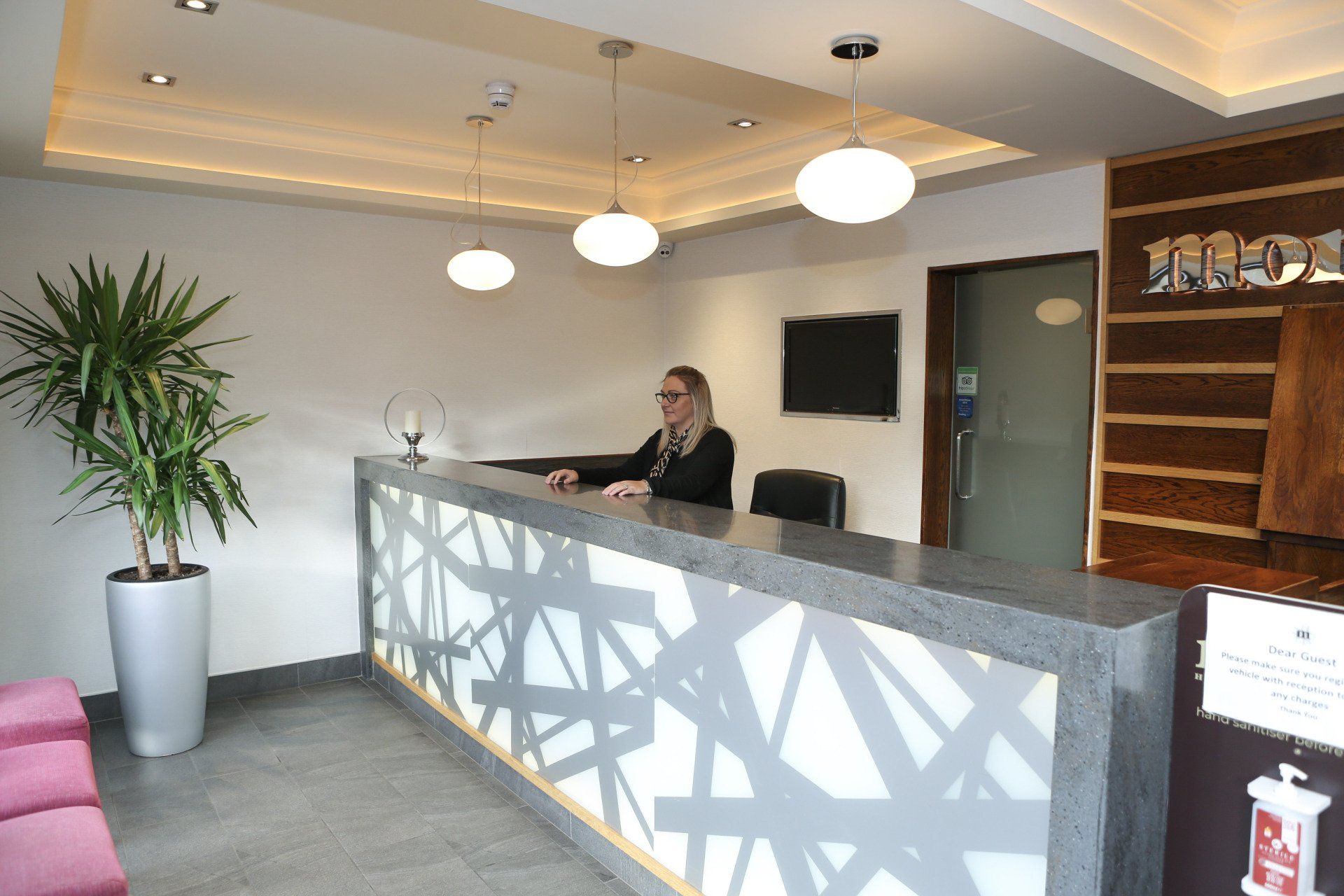 Hotel reception desk with woman receptionist. White, gray, and brown decor. Bright lights.