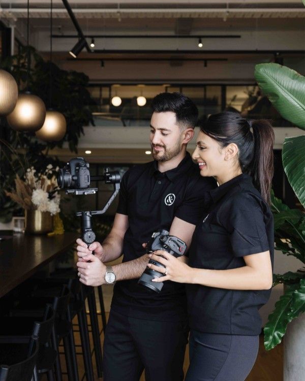 Ashika & Nathan, Clarke & Clarke, filming; both wearing black shirts, smiling.