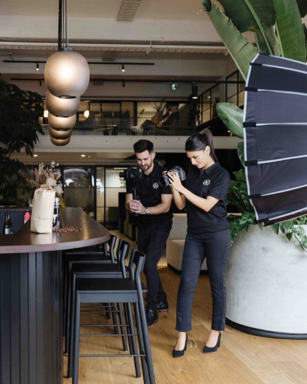 Two people, one holding a camera, preparing to photograph a bar area with a large light.