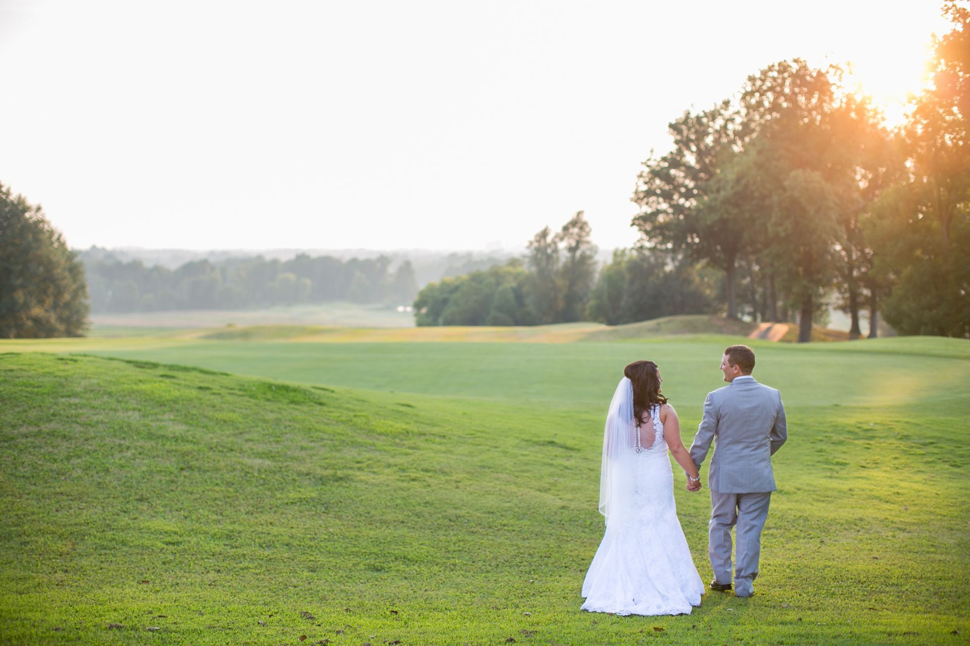 A bride and groom are walking through a grassy field holding hands.