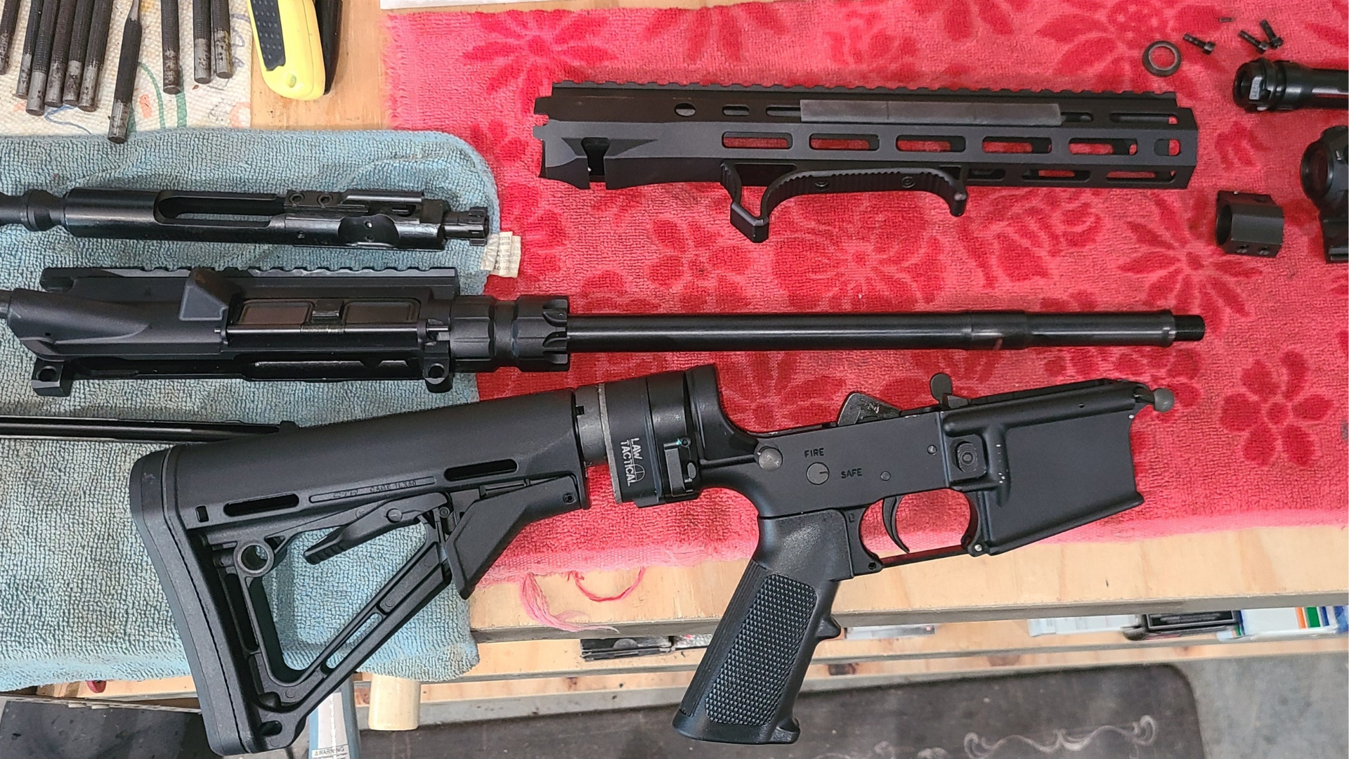 Several firearms and parts laid out on a red workbench, including black rifles and accessories.