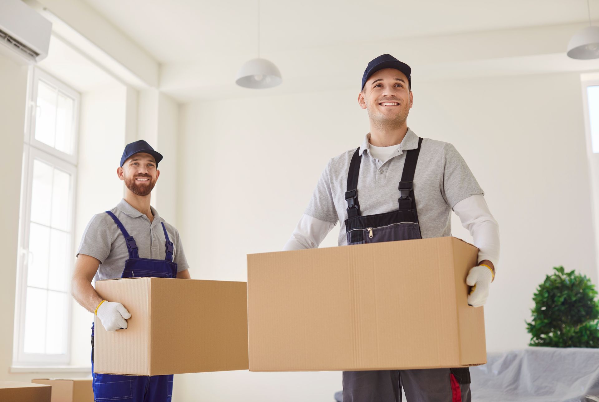 Two smiling movers in work uniforms carry cardboard boxes