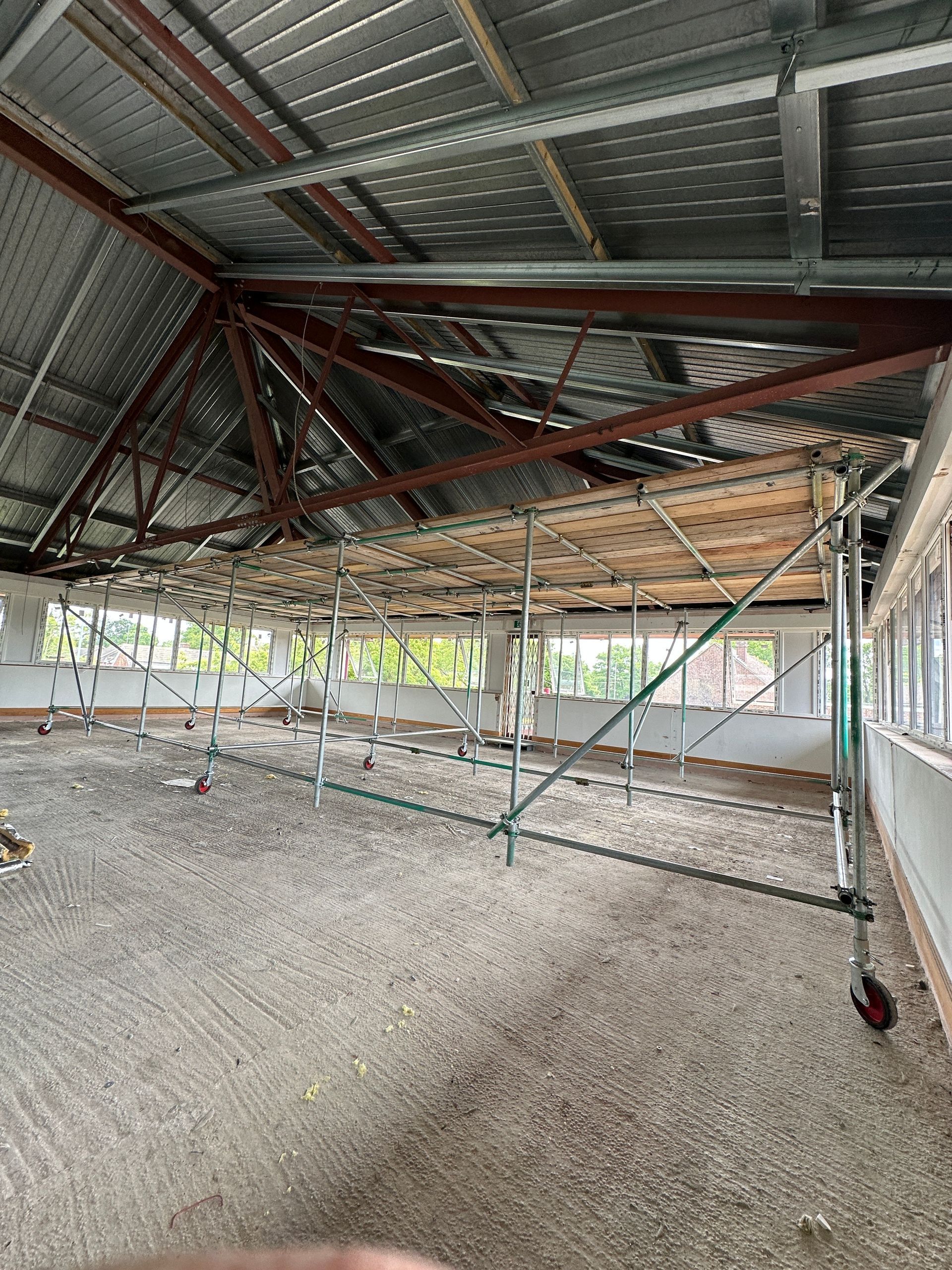 Interior view of a building under construction. Scaffolding supports wooden platforms under a metal roof. Concrete floor.
