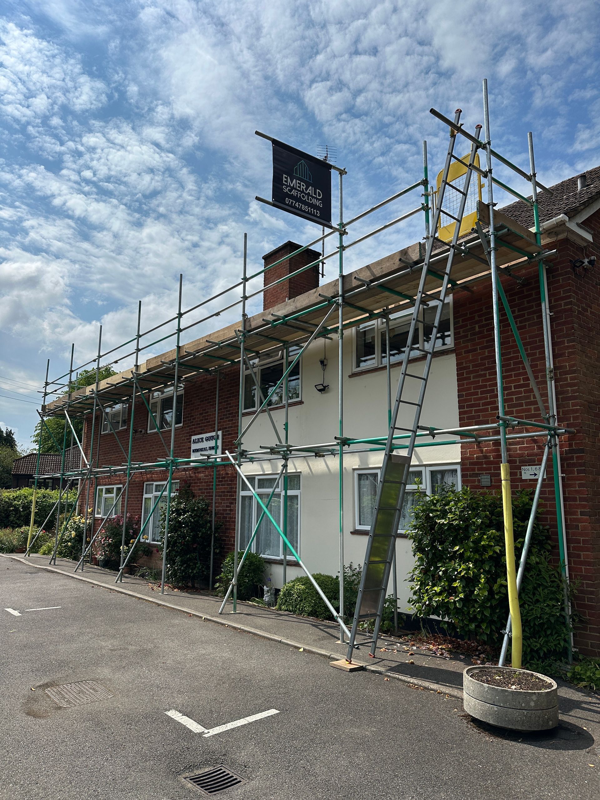 Scaffolding on a white and brick house, parked on a street, under a cloudy sky.