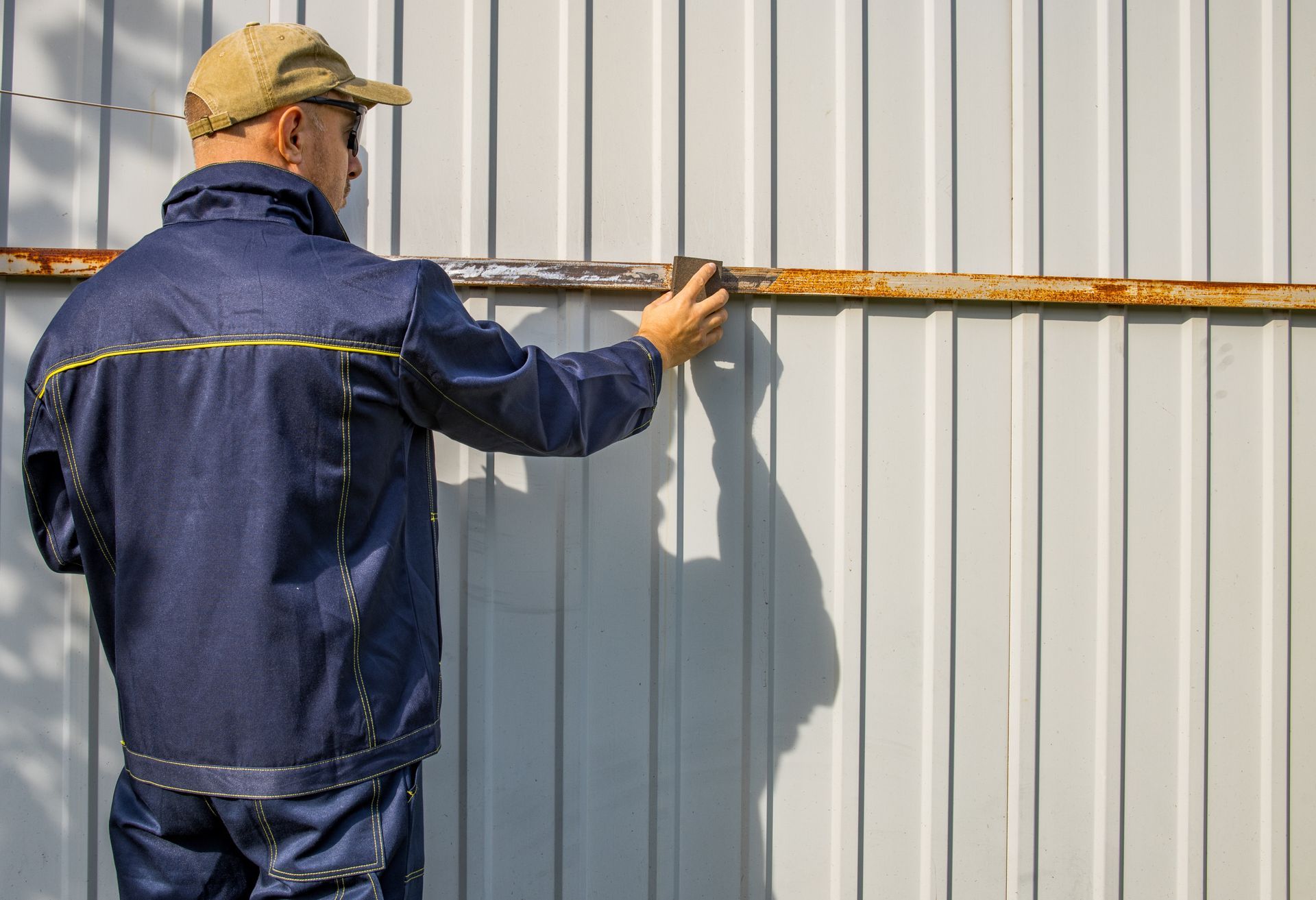 Worker in navy coveralls and cap touches a rusted rail on gray corrugated metal Worker in navy coveralls and cap touches a rusted rail on gray corrugated metal