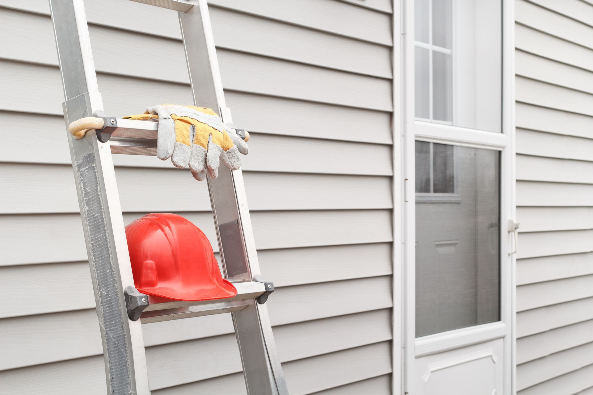 Ladder with gloves and hard hat leaning against house near door.