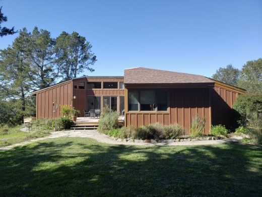 Brown house with multiple levels, surrounded by green grass and trees under a blue sky.