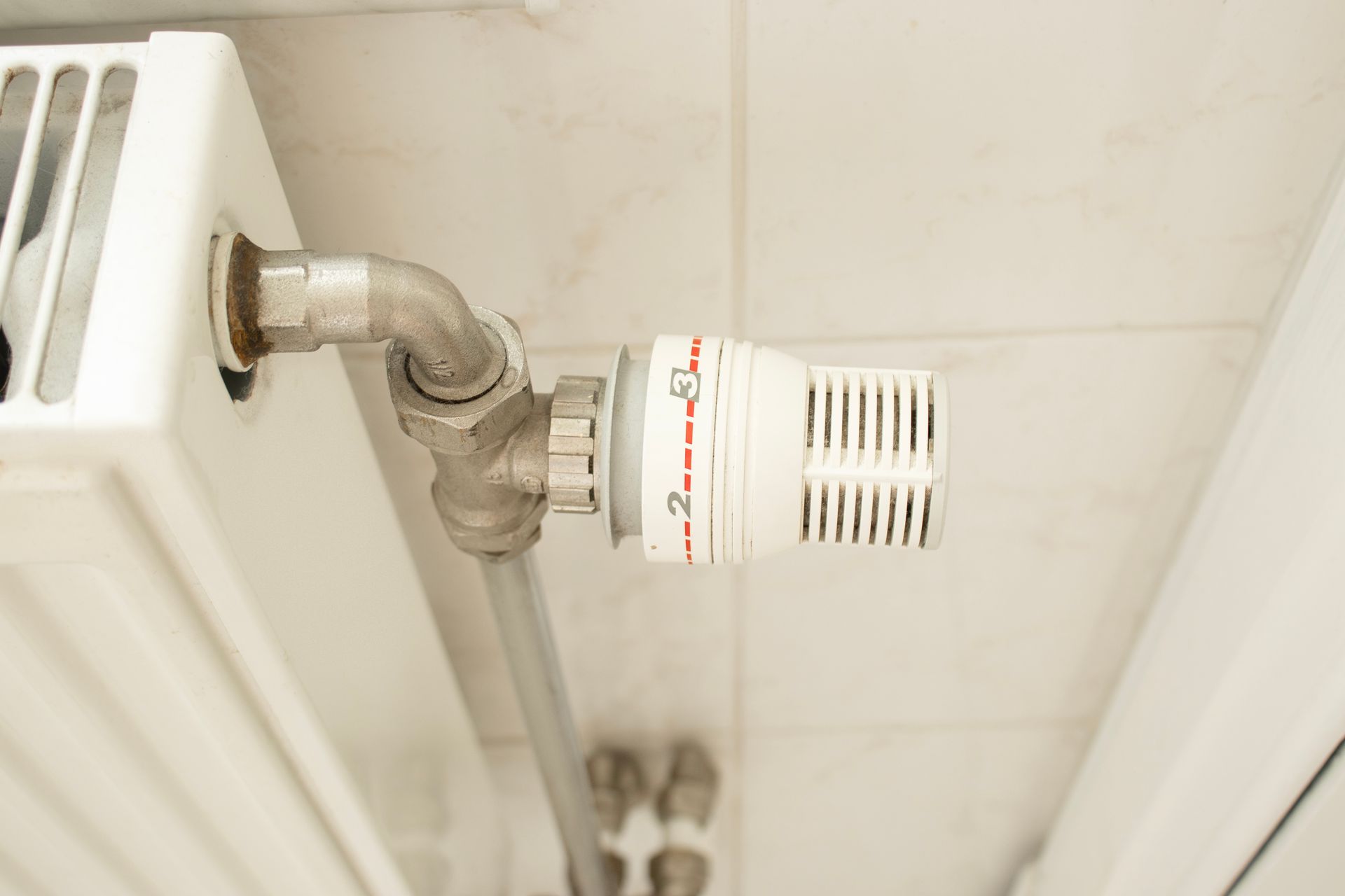 A white radiator with a white thermostatic valve set between numbers 2 and 3, mounted against a white tiled wall. A white radiator with a white thermostatic valve set between numbers 2 and 3, mounted against a white tiled wall.
