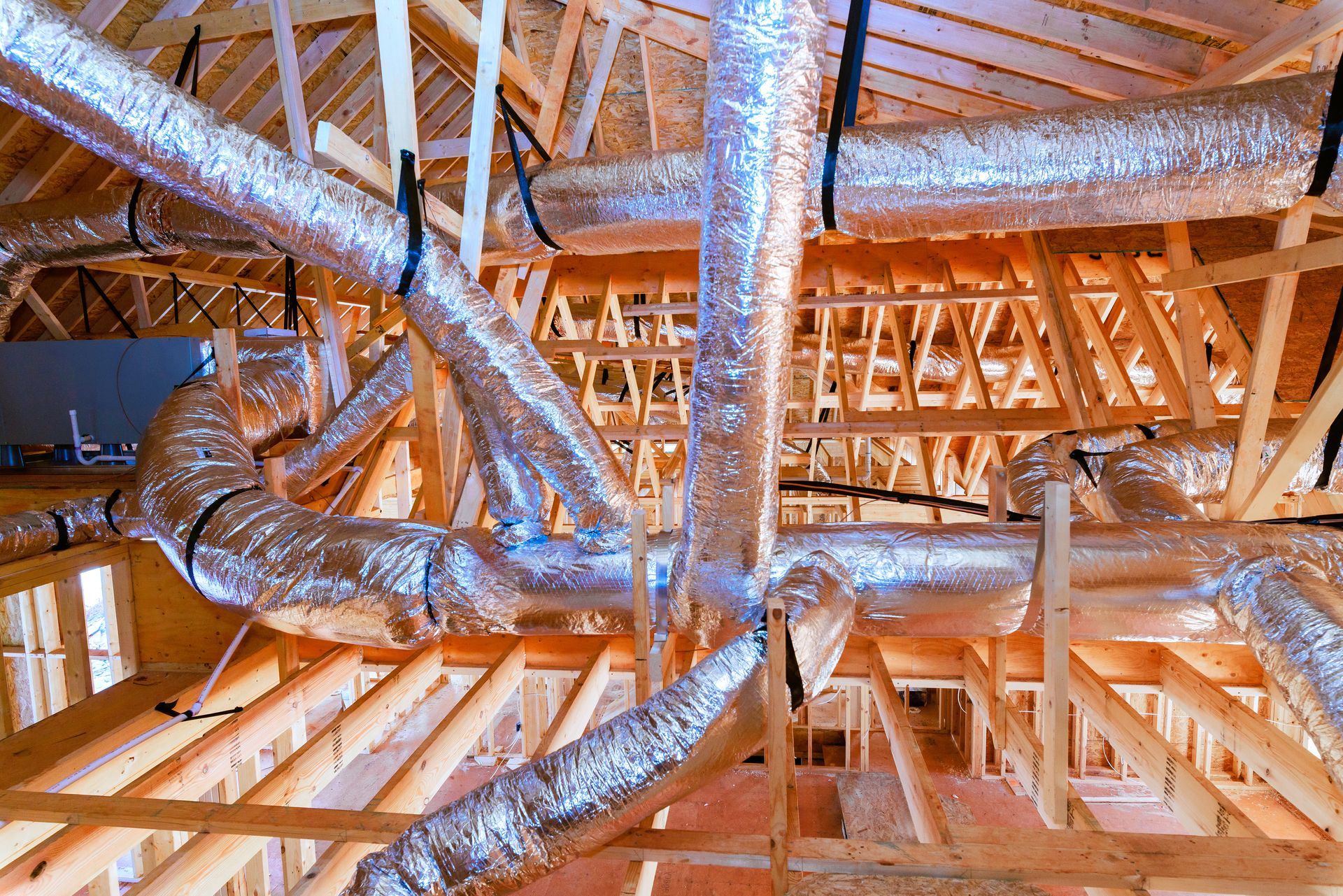 Insulated silver HVAC ducting installed throughout the wooden attic framework of a building under construction. Insulated silver HVAC ducting installed throughout the wooden attic framework of a building under construction.