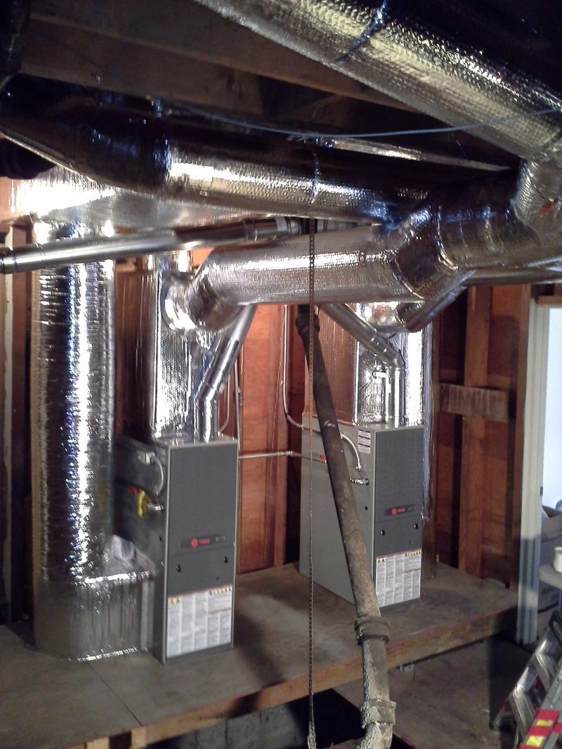 Metal HVAC ductwork and furnace units in an unfinished basement, with insulated pipes overhead.